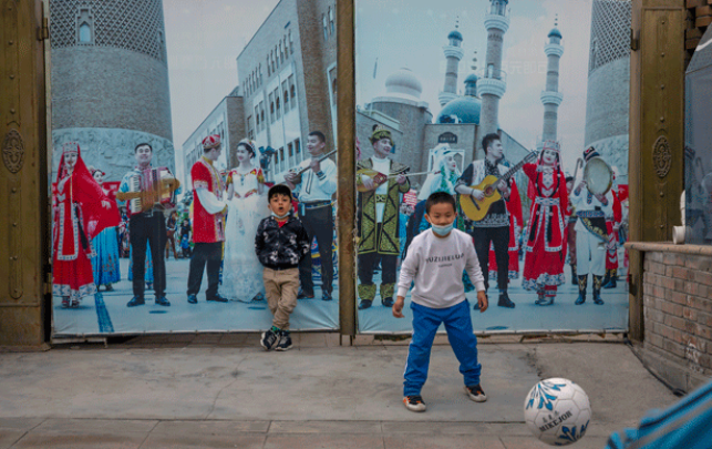 Anak-anak bermain bola di depan mural yang menunjukkan para musisi Uyghur tampil pada acara International Grand Bazaar di Urumqi, ibu kota Xinjiang. Foto diambil pada saat kunjungan wartawan internasional tanggal 21 April 2021, yang diorganisir oleh rezim komunis Cina. Foto: AP 